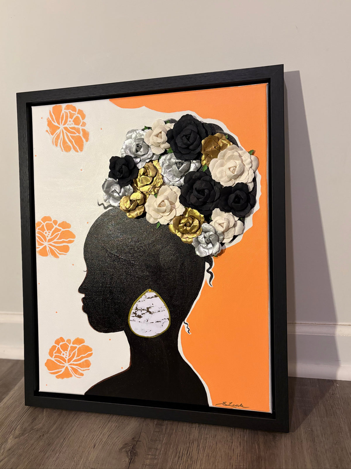 A black framed art painting featuring a black silhouette of a woman in profile with large floral hair that is white, black, and gold, set against a split cream and orange background. 